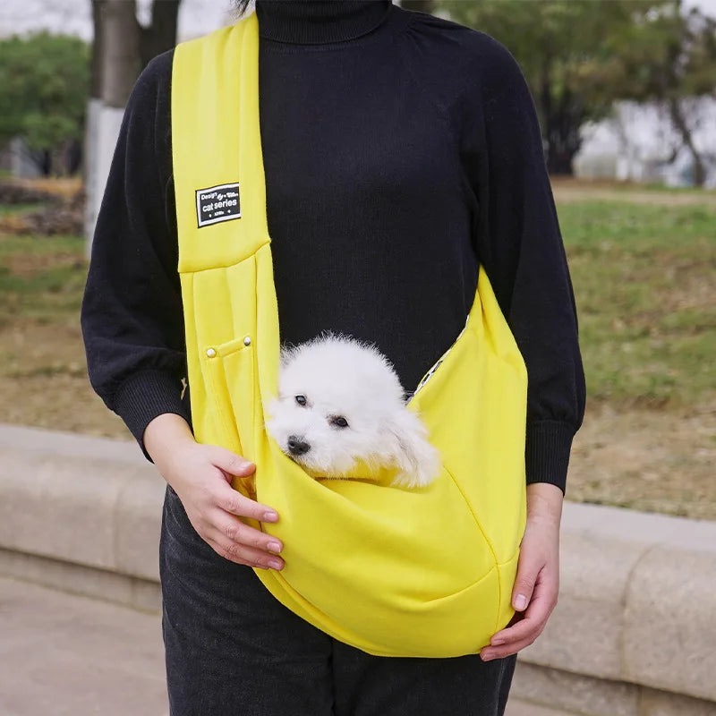 A person is holding a bright yellow portable pet carrier sling bag with a white puppy inside, against a blurred outdoor backg