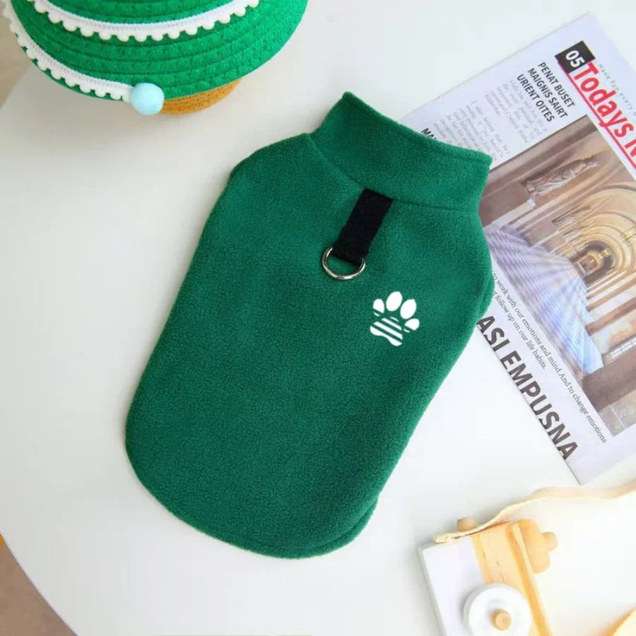 A dark green fleece dog sweater with a black tag and a white paw print embroidery is displayed next to a green knitted hat.