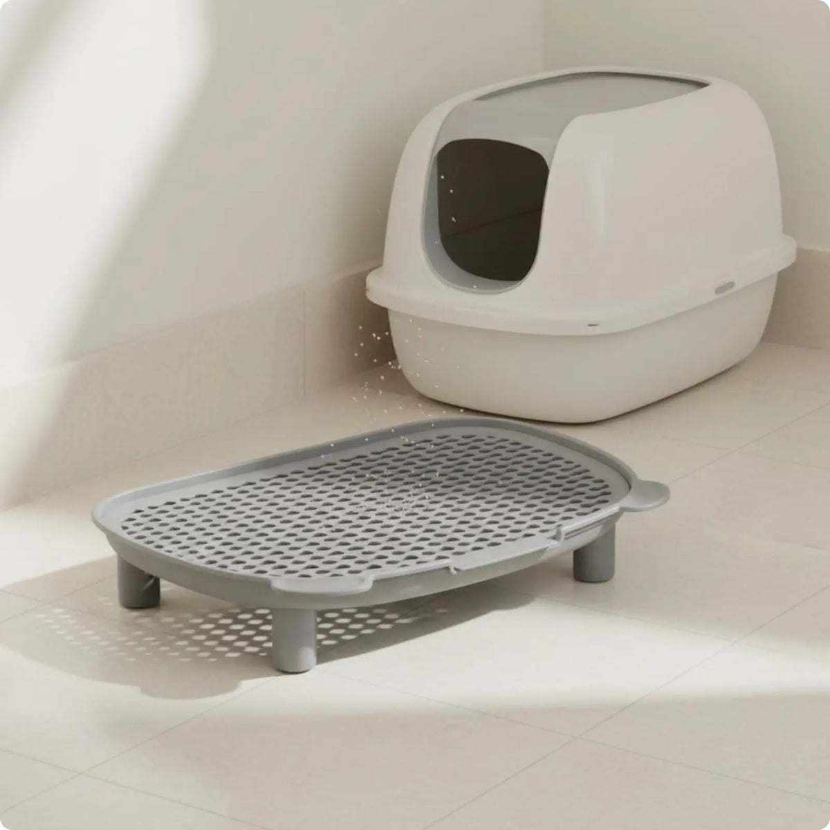 A white smart cat litter box sits on a floor next to a gray, perforated litter tray.