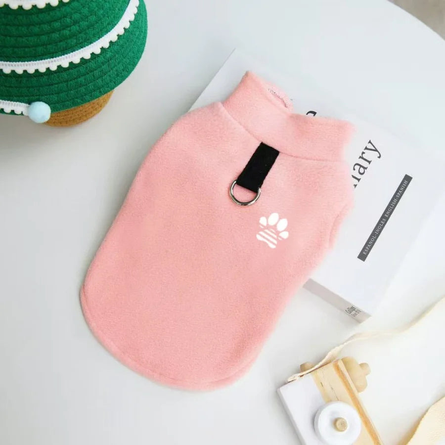 A pink cozy fleece dog sweater with a black tag is displayed on a white surface next to a green hat and a book.
