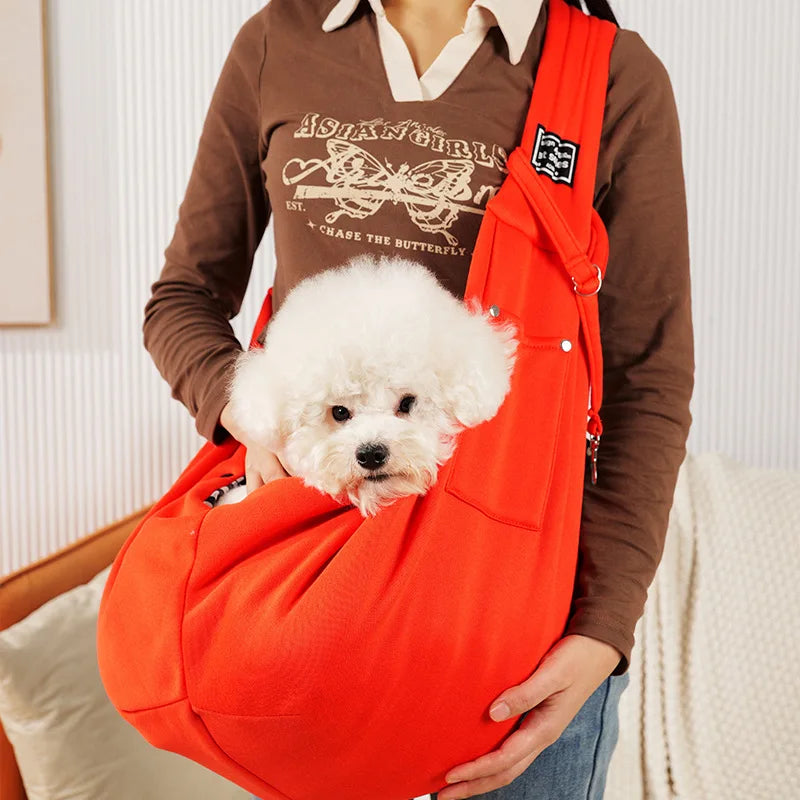 A person holds an orange portable pet carrier sling bag containing a white fluffy dog.