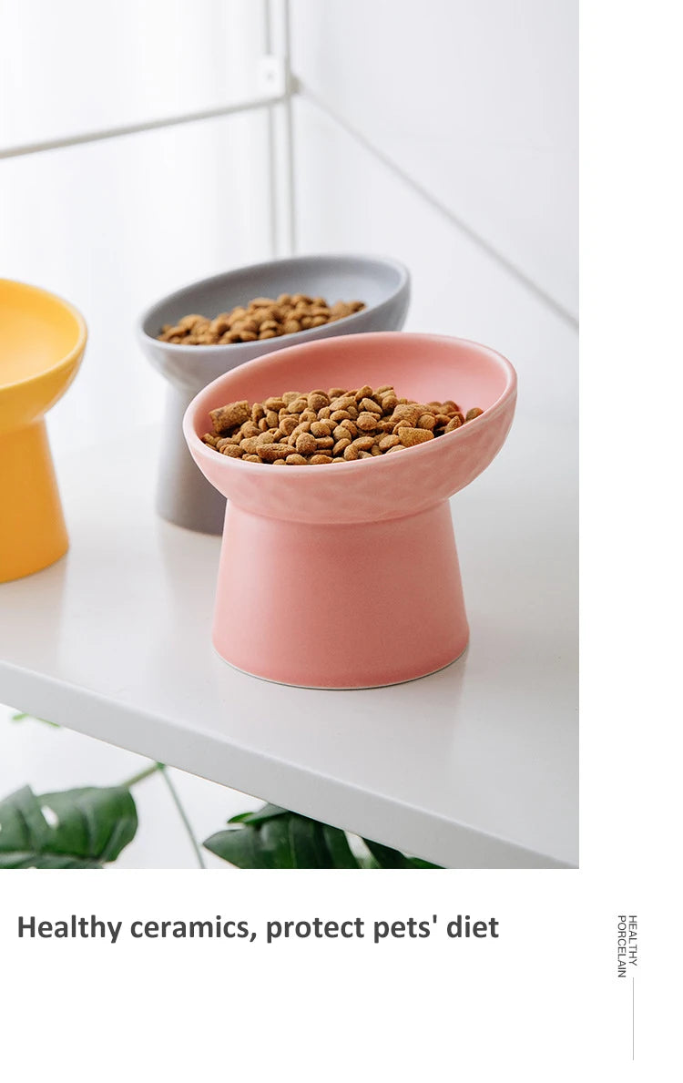 A pink ceramic food bowl filled with dry pet food sits on a white surface next to a gray bowl.