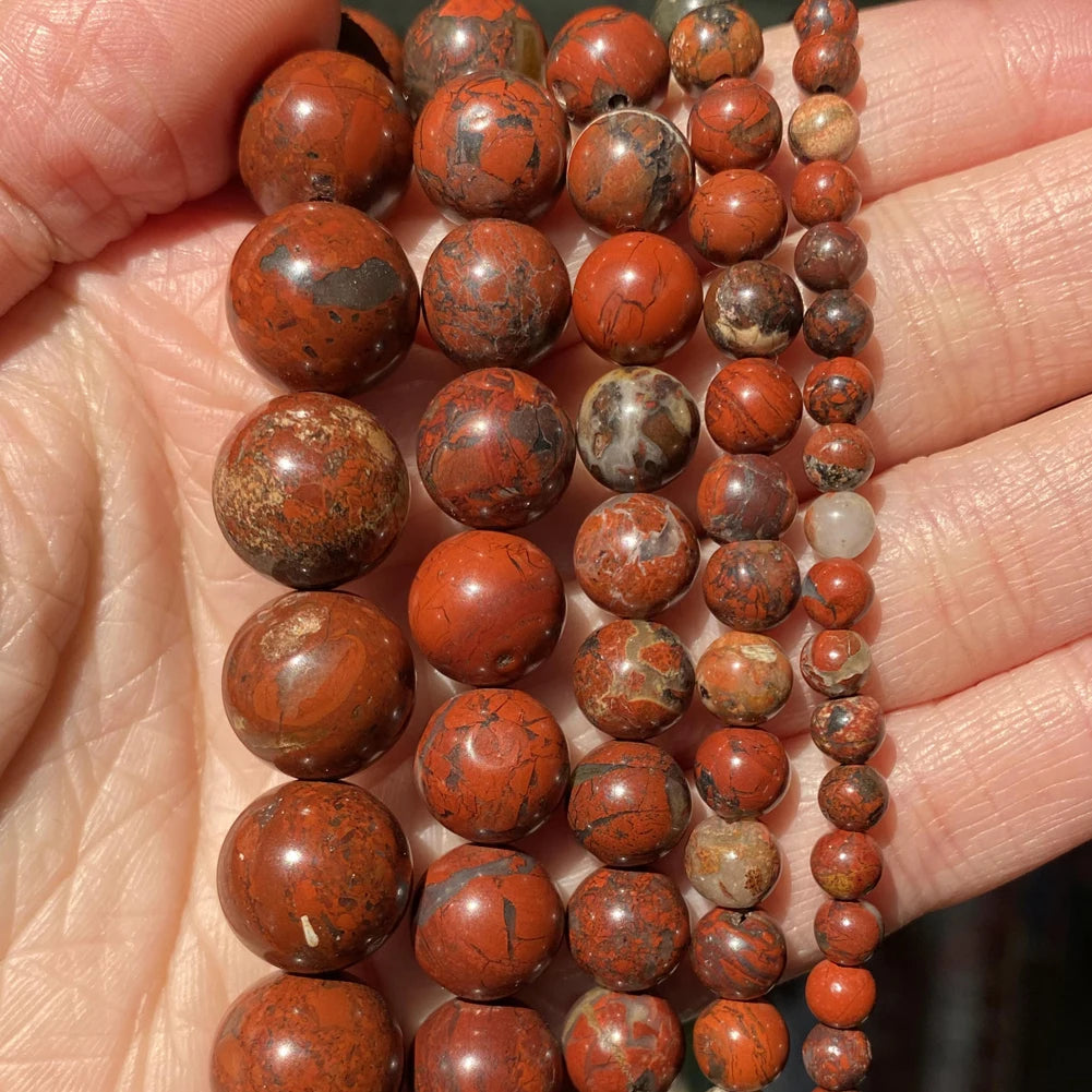 A hand holds a string of natural reddish-brown stone beads for jewelry making in various sizes and styles.