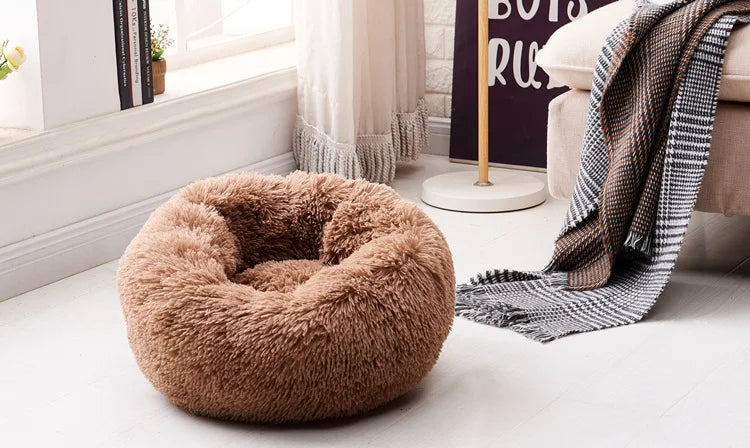 A plush, round, brown pet bed with a textured fur cover resting on a light wooden floor.