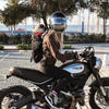 A woman is riding a motorcycle with a dog in a backpack carrier outdoors.