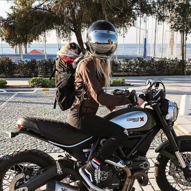 A woman is riding a motorcycle with a dog in a backpack carrier outdoors.