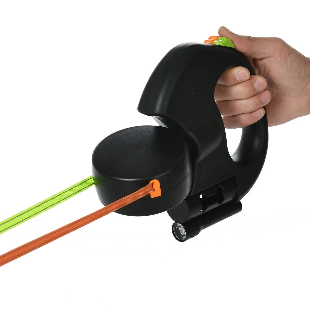A hand is holding a black retractable dual dog leash with an orange reflective nylon strap.