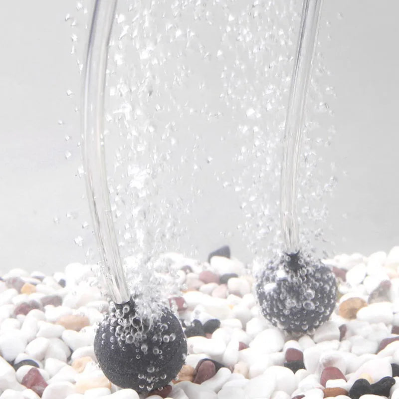 A close-up shot of five black round aquarium air stones being vigorously blown by water spray onto a white gravel surface.