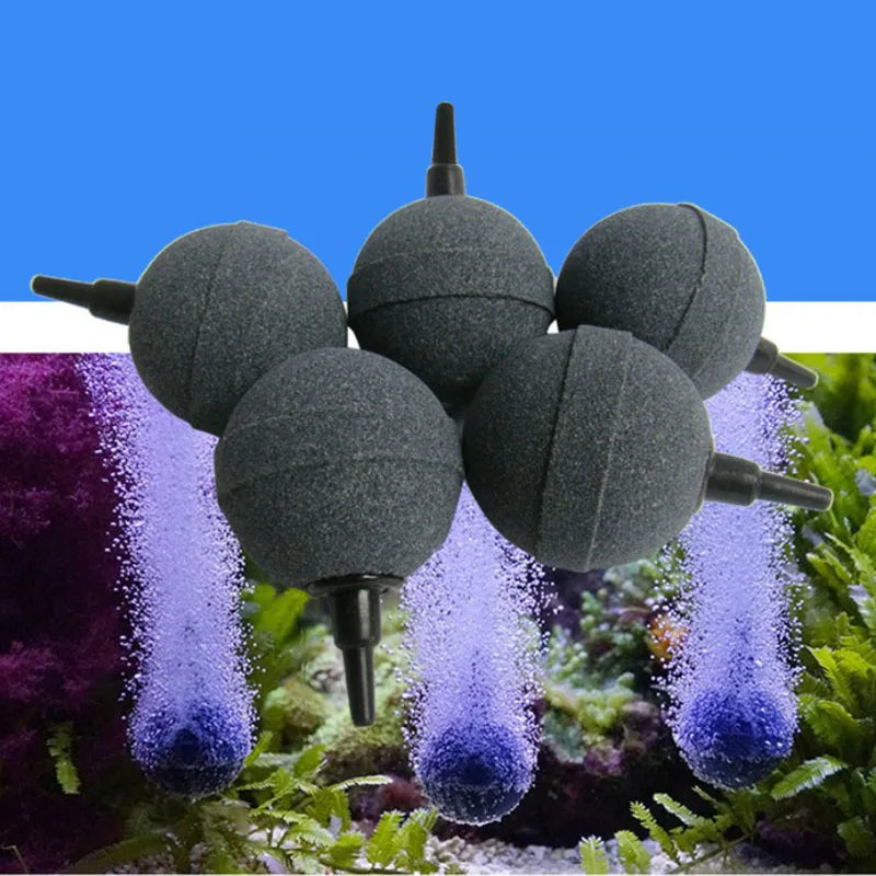 A set of five dark gray mini round aquarium air stones are shown against a blurred background of green aquatic plants.