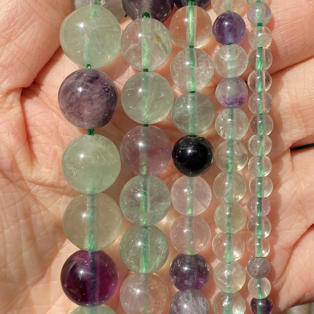 A hand holds a string of natural stone beads in various shades of green, purple, and pink.