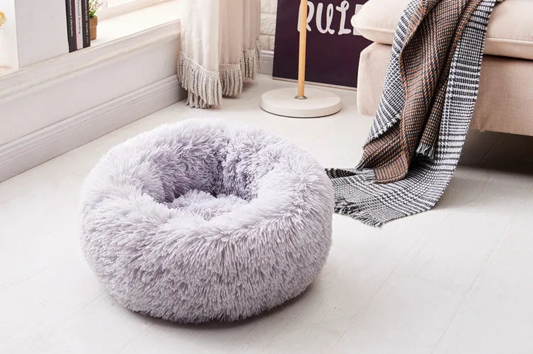 A fluffy, round pet bed with a gray faux fur exterior and anti-slip base resting on a light wooden floor.
