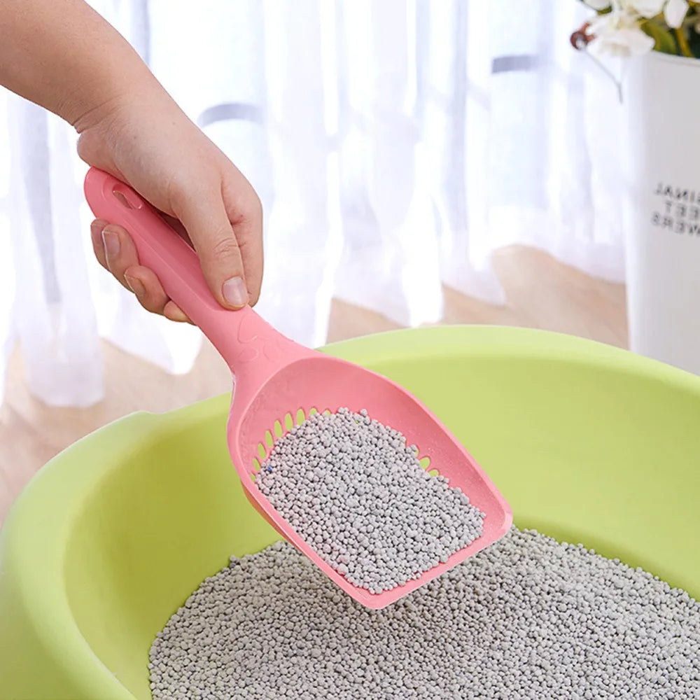 A hand is using a pink pet litter scoop with a hollow sifting head to transfer gray pet litter into a green bowl.