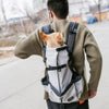 A person is wearing a beige sweater and carrying a white breathable dog backpack carrier with an orange tag on their back.