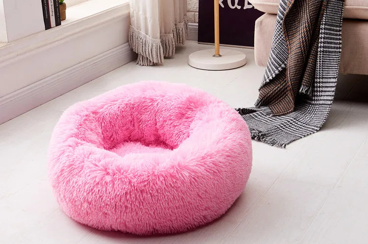 A fluffy pink round pet bed with an anti-slip base resting on a light wooden floor.