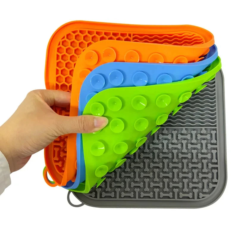 A hand holds a stack of brightly colored silicone pet licking mats with suction cups on a gray feeding mat.
