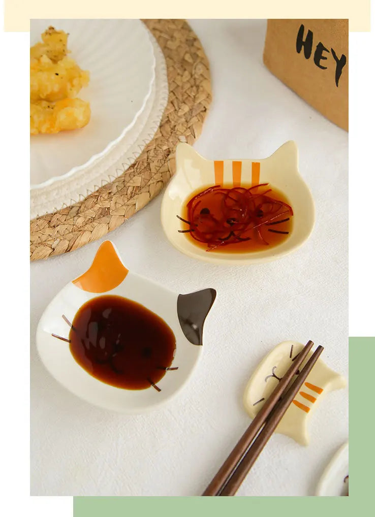 A ceramic cat-shaped sauce dish holds dark brown liquid, accompanied by chopsticks and a piece of food on a white plate.