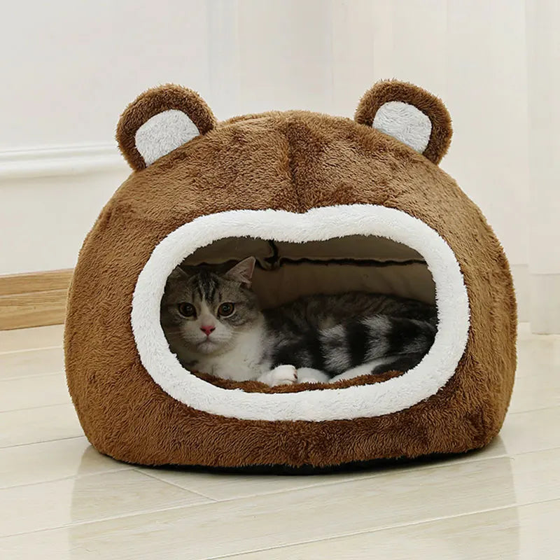 Un lit de grotte pour chat en peluche marron avec de grandes oreilles et une ouverture où se repose un chaton tabby.