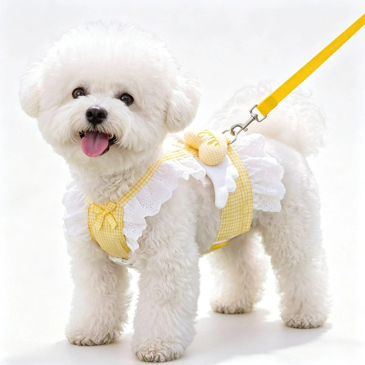 A fluffy white dog wearing a yellow plaid harness vest with white lace ruffles and an angel heart design.
