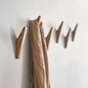 A light tan coat hangs on an oak wood wall mounted triangle coat rack with three hooks against a white wall.