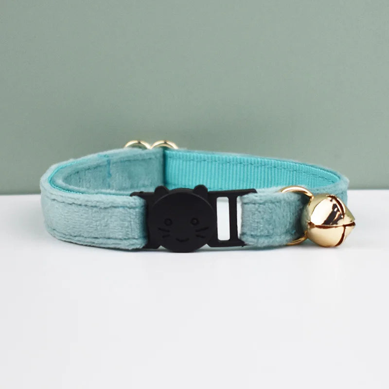 A light blue velvet cat collar with a black cat charm and a gold safety buckle is displayed on a white surface.