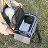 A person is holding an open gray expandable pet carrier backpack with black mesh and reflective trim on a grassy field.