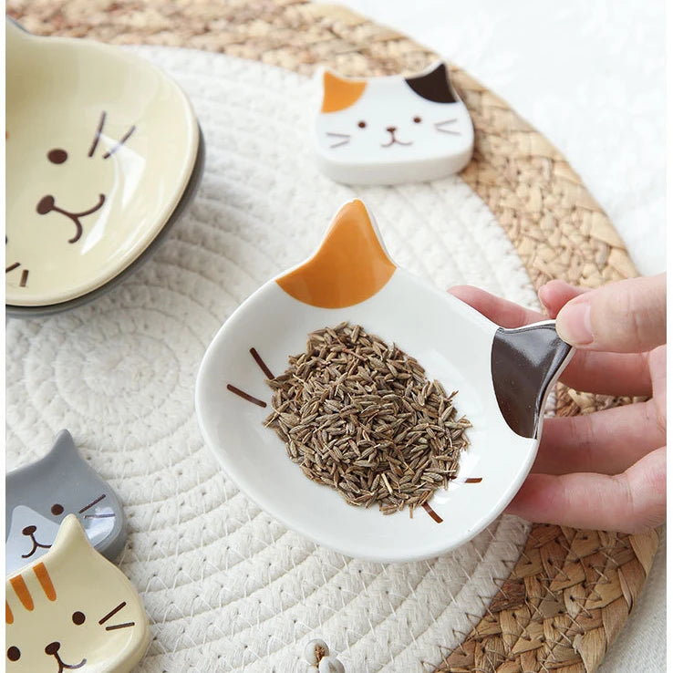 A hand holds a white ceramic cat-shaped sauce dish filled with dried herbs on a woven mat.