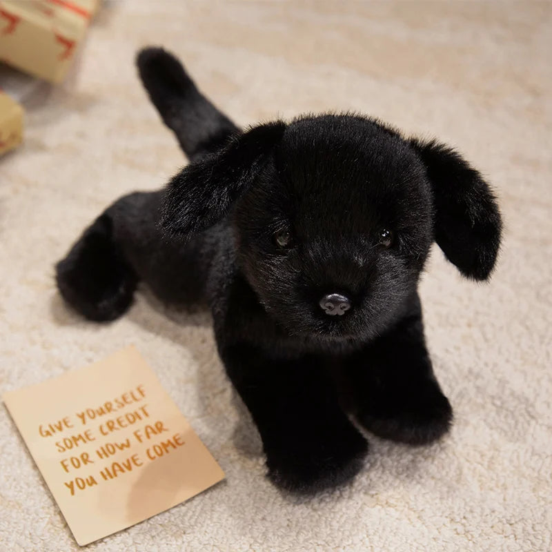 A black realistic Labrador plush toy lying on a light-colored surface next to a note.