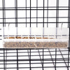 A clear plastic hanging bird feeder and waterer with a splash guard is suspended in a black wire grid.
