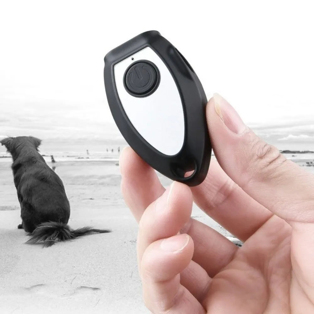 A hand holds a black and white handheld ultrasonic dog bark deterrent device against a gray beach background with a dog nearb