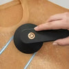 A hand is operating a black portable electric lint and fabric shaver with an LED display on a light wood surface.