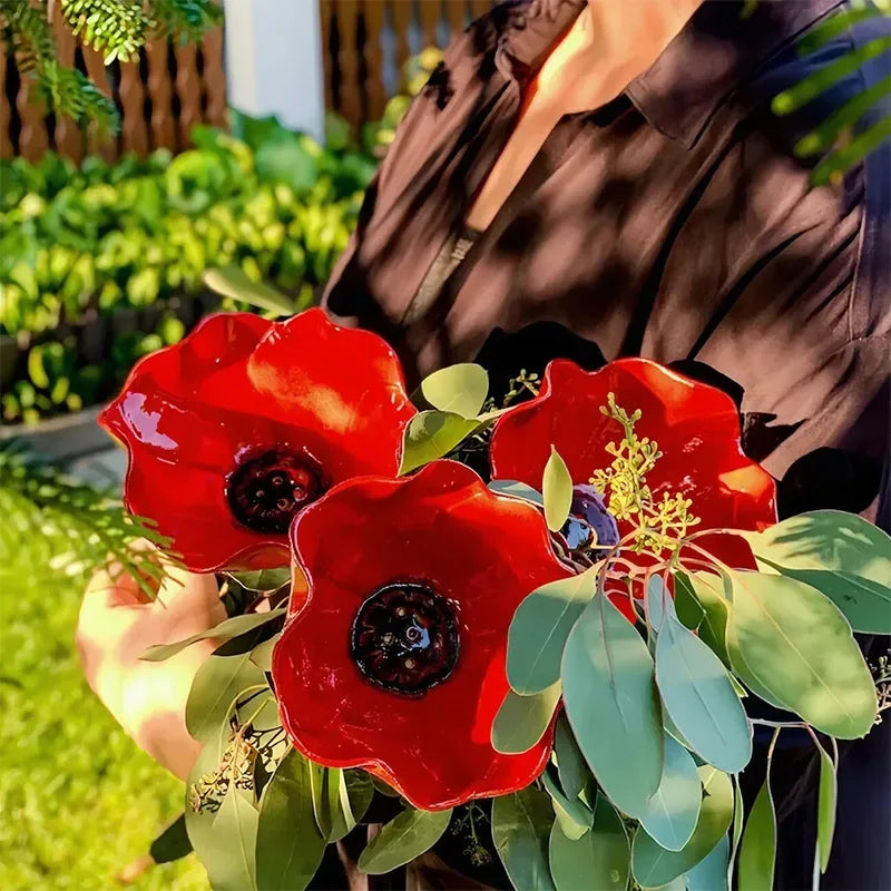 A hand holds a vibrant red flower-shaped outdoor bird bath with green leaves in a garden setting.