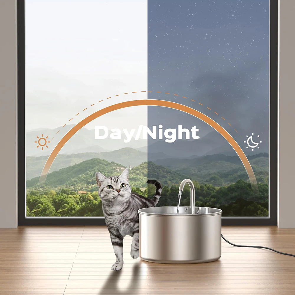 A stainless steel pet water fountain is placed on a wooden floor in front of a window displaying a day/night landscape.