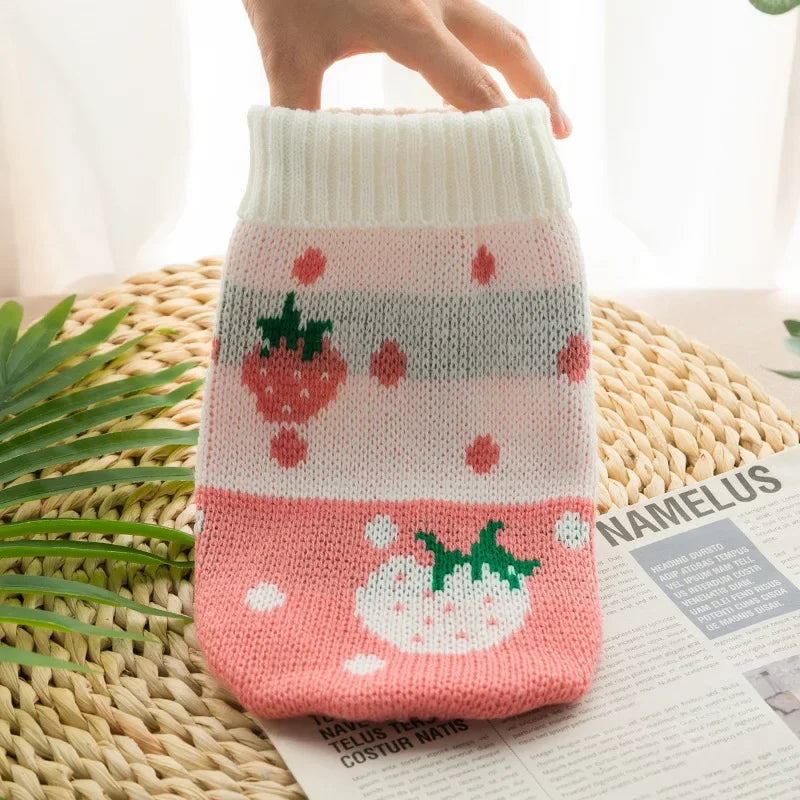 A pink knitted dog sweater with red strawberry patterns rests on a woven mat next to some papers.