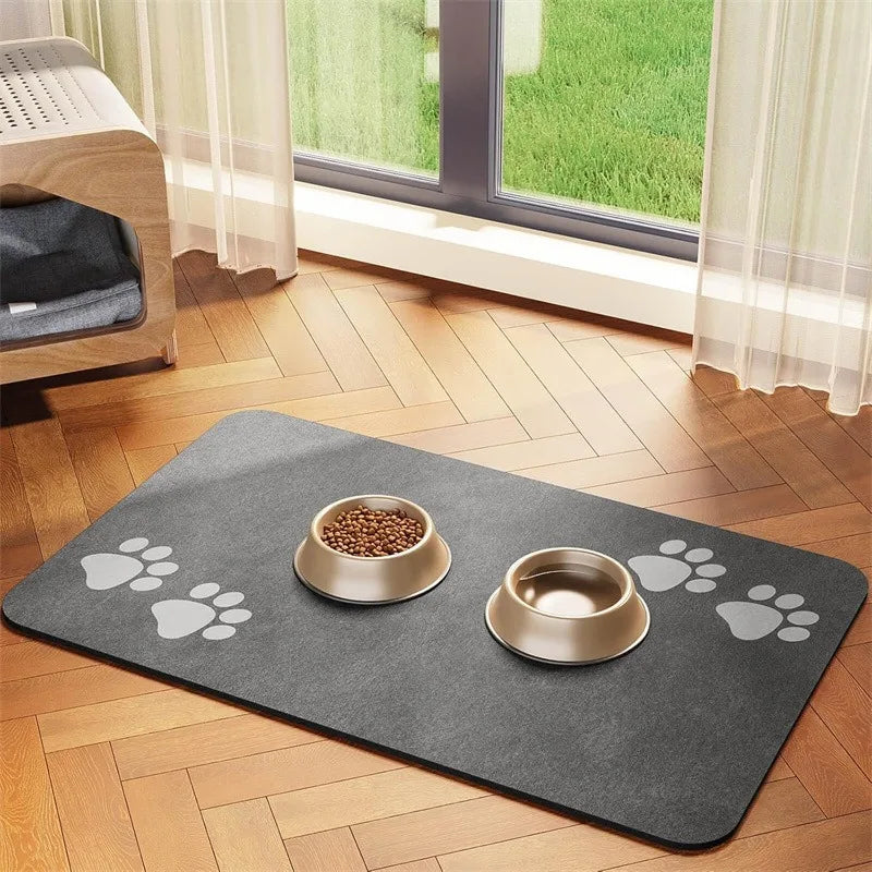 A gray non-slip pet feeding mat with paw prints and bowls placed on a wooden floor near a window.
