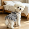 A fluffy white dog wearing a black and white checkered ruffled tank top sitting on a wooden floor.