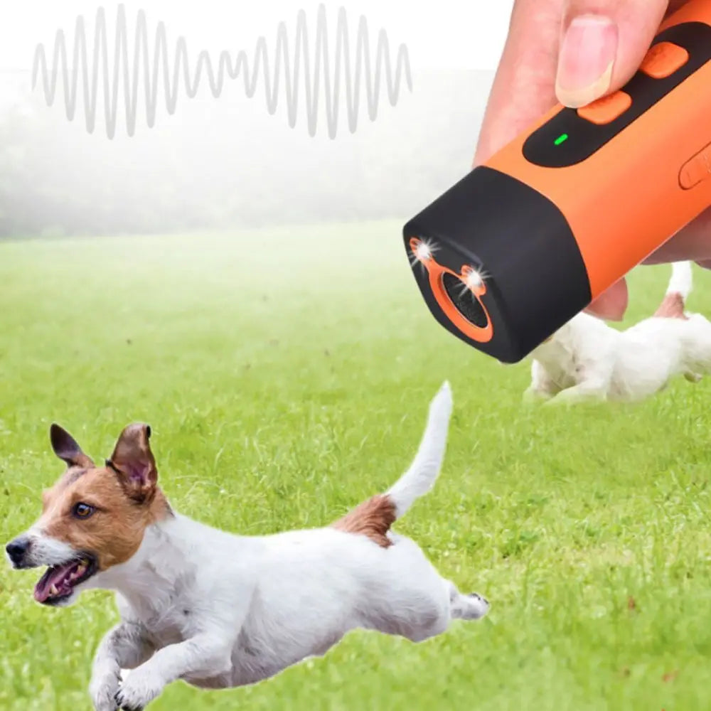 A hand holds an orange ultrasonic dog repeller device while a dog runs past in green grass.