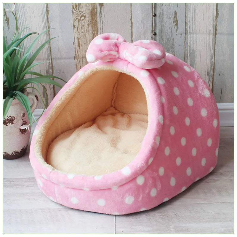A pink, polka-dotted cozy cave pet bed with a fluffy interior is shown on a light wooden surface.