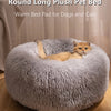 A round, long gray plush pet bed with a cat resting inside on a wooden floor.