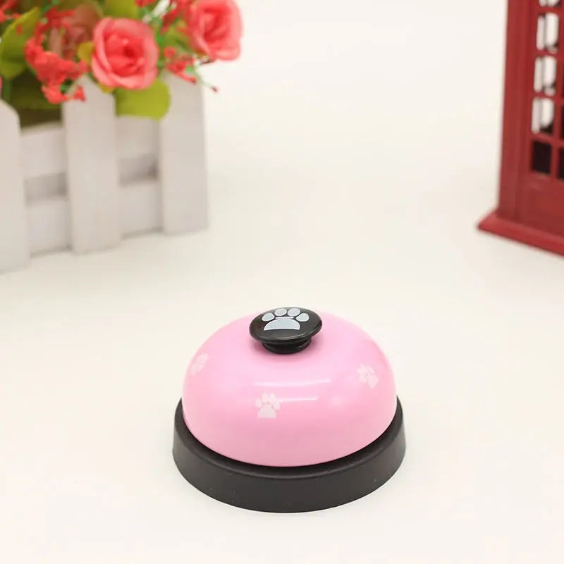 A pink and black footprint pet training bell resting on a white surface with blurred flowers in the background.