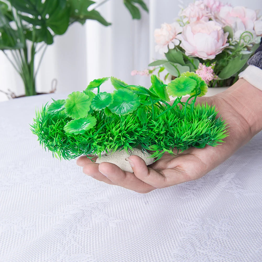 Una mano sostiene una vibrante planta acuática artificial verde con una base de cerámica sobre una superficie blanca.