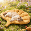 A fluffy white cat is sleeping on a yellow leaf-print pet cushion outdoors in a grassy area.