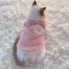 A fluffy white cat wearing a pink embroidered vest on a white, textured surface.