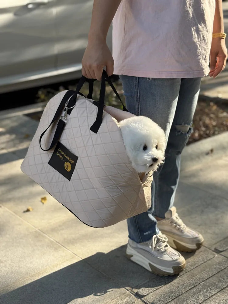 A person is holding a light pink quilted pet carrier bag with black handles and a white fluffy dog inside, standing outdoors.