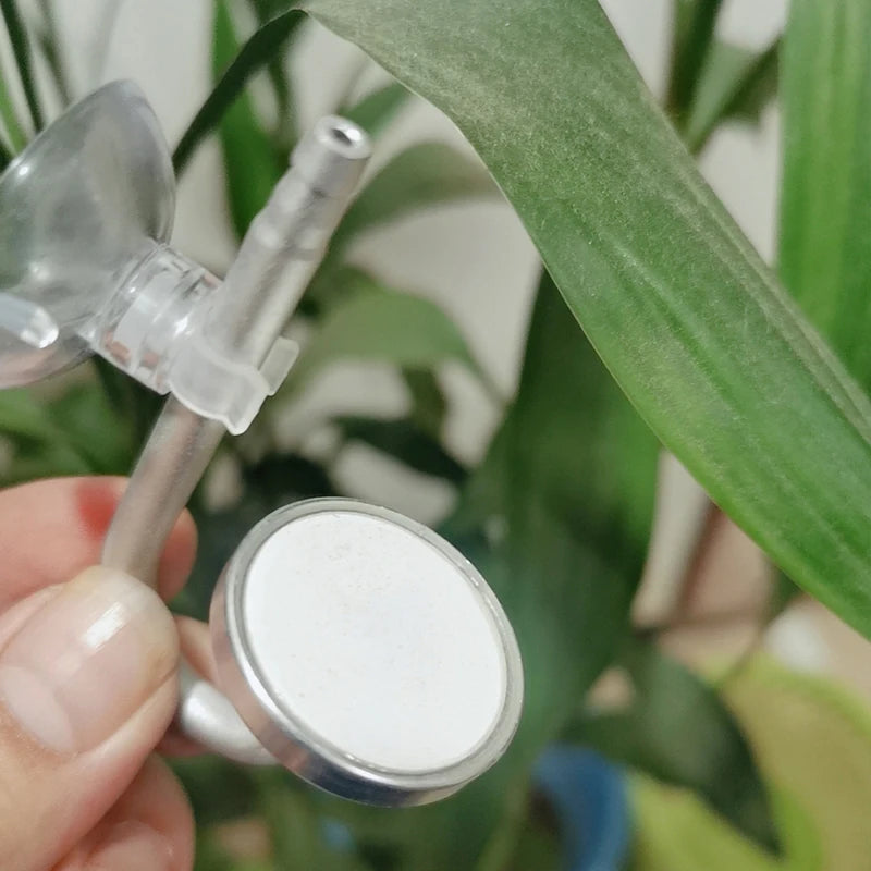 Eine Hand hält einen Aluminium-CO2-Diffusor, ein kleines silbernes Gerät mit einer weißen Scheibe, neben grünem Laub.