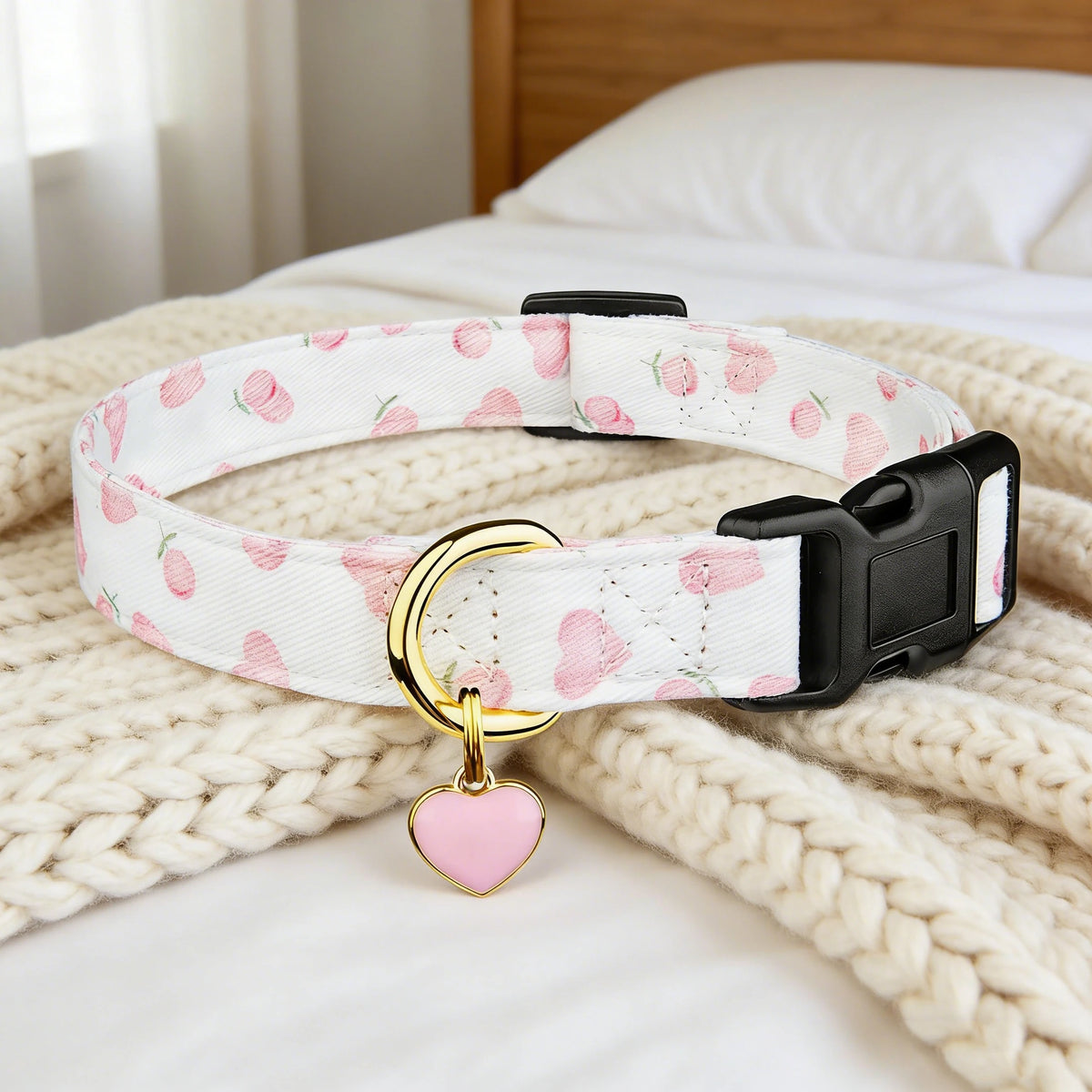 Rosa und weiß gemusterter Baumwollhundkragen mit goldenem Herz- und Erdbeere-Anhänger auf cremefarbener Strickdecke.