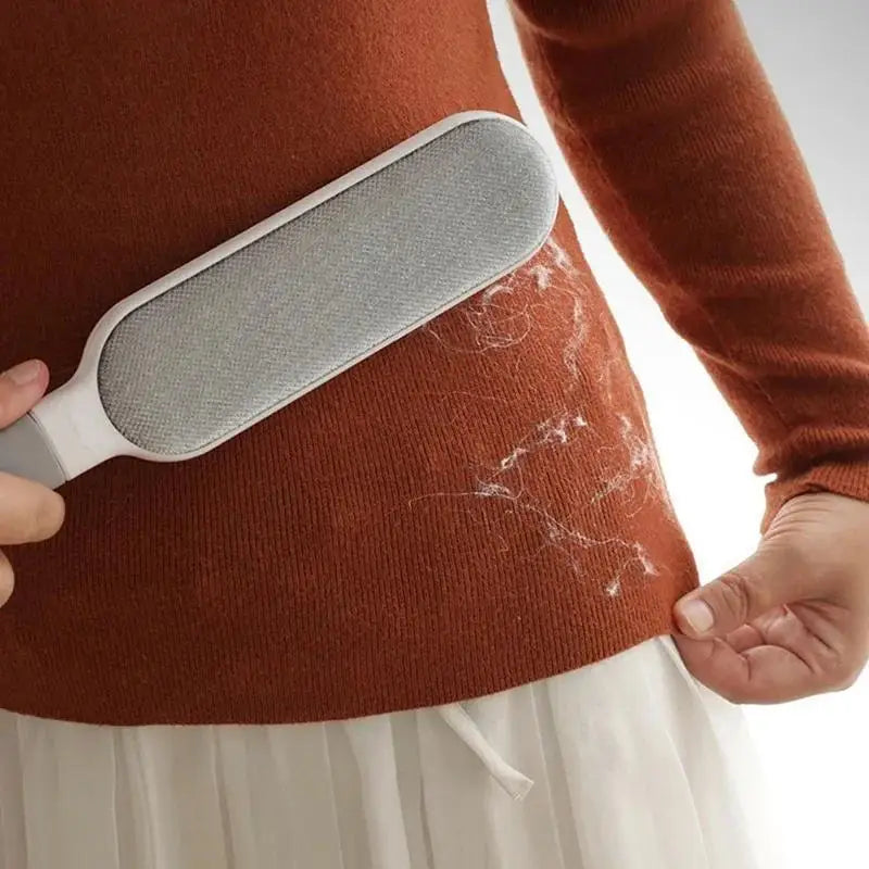 Une main tient une brosse griffe à poils pour animaux grise contre le pull brun-rouge d'une femme.