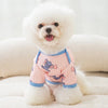 A white dog wearing a pink cartoon print vest is lying down on a light surface.