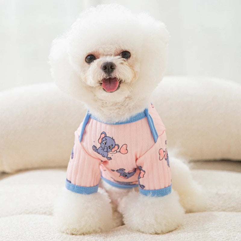 A white dog wearing a pink cartoon print vest is lying down on a light surface.