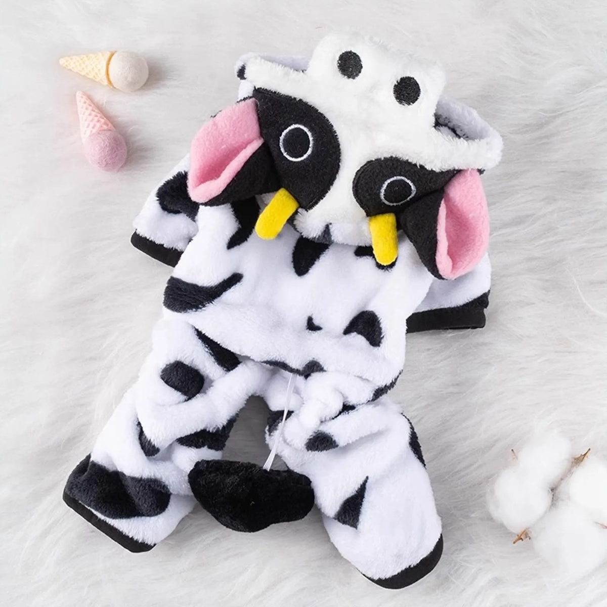 A white and black cow print dog costume with a hood is displayed on a fluffy white surface.