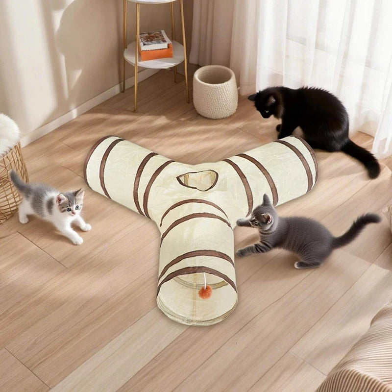 A beige and brown foldable pet play tunnel for cats is lying on a wooden floor with several cats nearby.